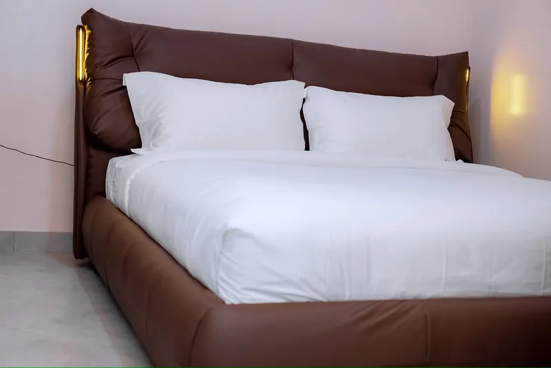 Furnished bed with white linen, brown headboard and warm bedside lighting